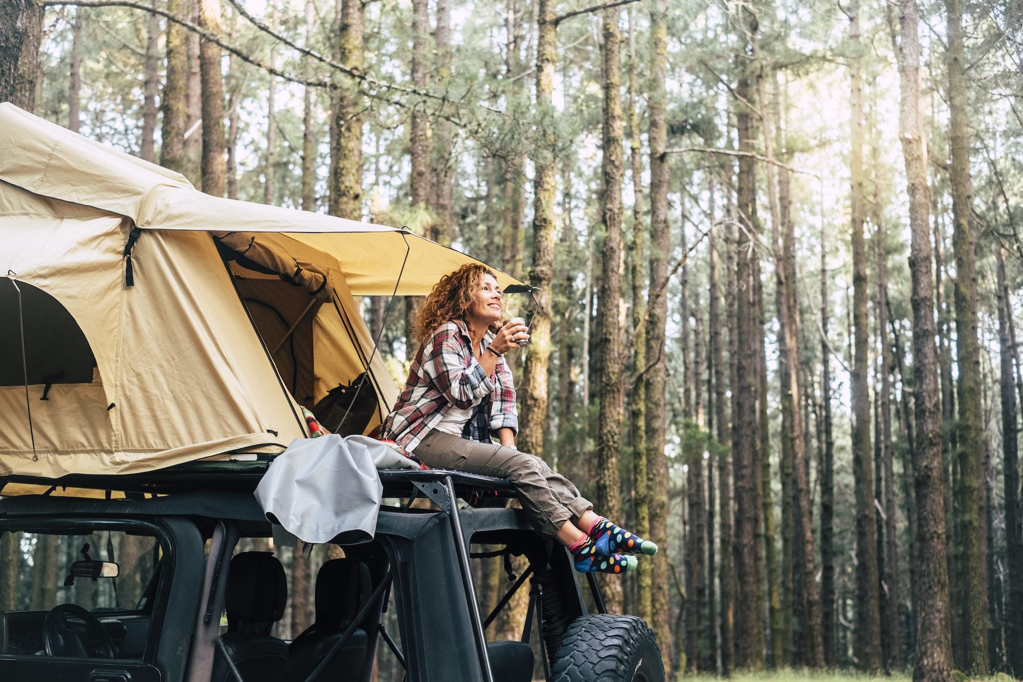 Young woman sitting outside tent on the roof of the car and drinking coffee in forest. Woman relax